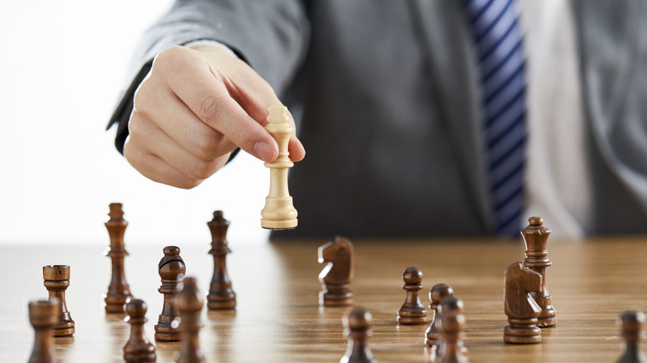 Businessman In A Suit Using His White King Chess Piece Among Dark Chess Pieces On A Table Businessman In A Suit Using His White King Chess Piece Among Dark Chess Pieces On A Table