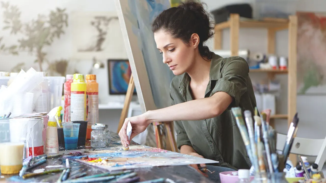 Serious Brunette Young Beautiful Woman Sitting In Art Studio, Taking Colorful Paints From Tube While Creating Great Masterpiece On Easel, Being Preoccupied With Her Work, Having Nice Imagination