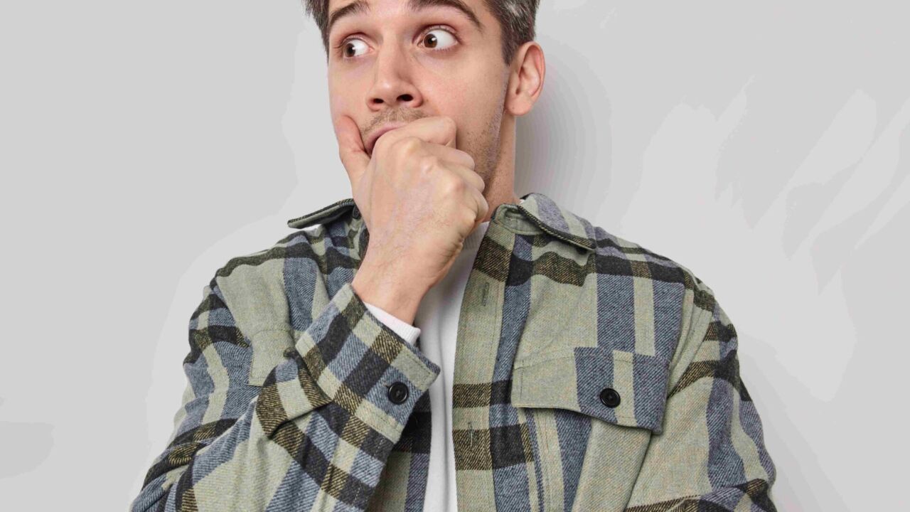 Shocked Young European Adult Man Stares Bugged Eyes Covers Mouth With Hand Wears Checkered Shirt Concentrated Away Blank Copy Space For Your Advertisement. Human Reactions And Emotions Concept